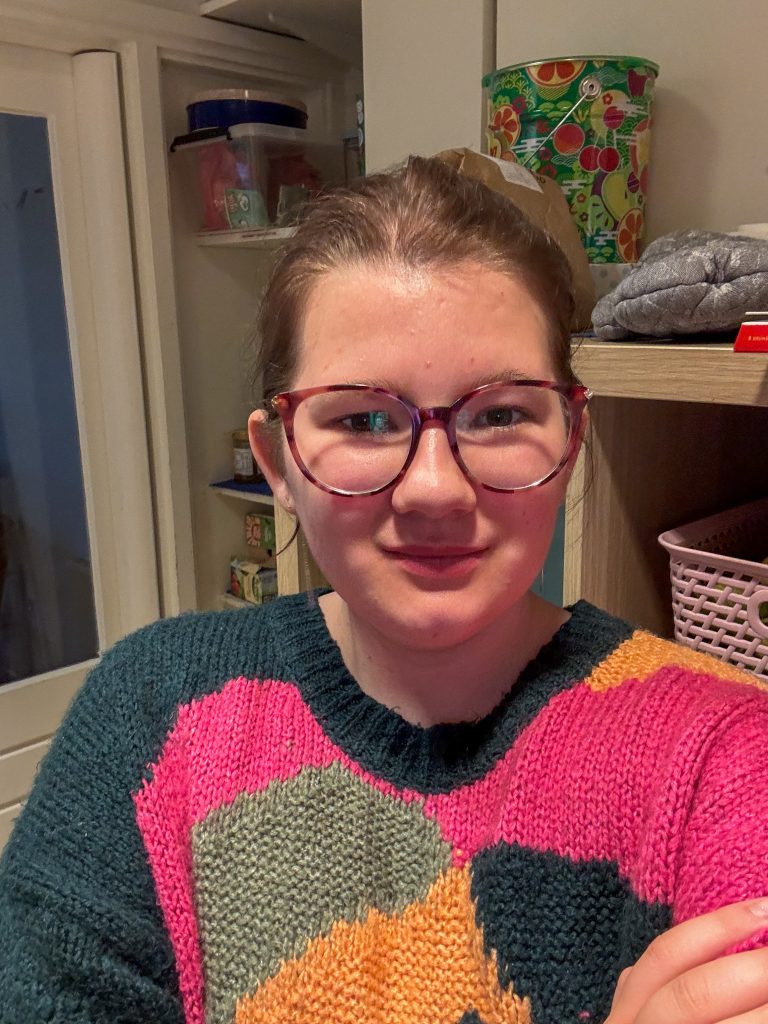 Beaudine met een kleurrijke gebreide trui en bril, lachend in de keuken op haar werk.