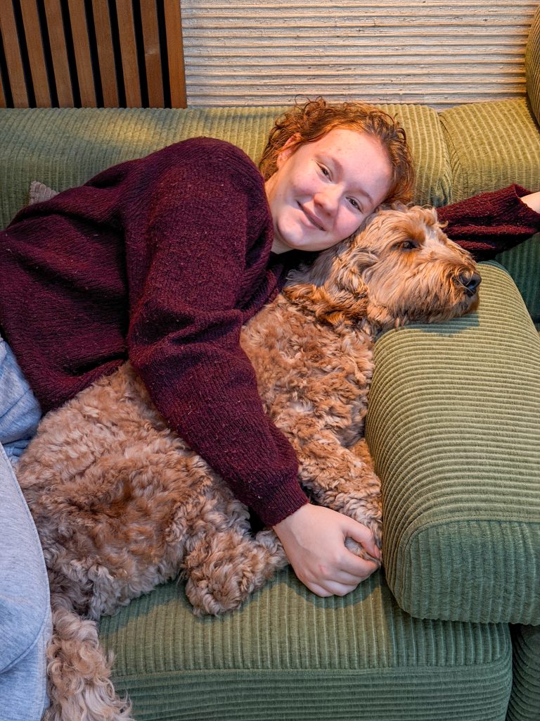 Amaranthe ligt op de groene bank en knuffelt met hond Teun tijdens een rustig moment binnen.