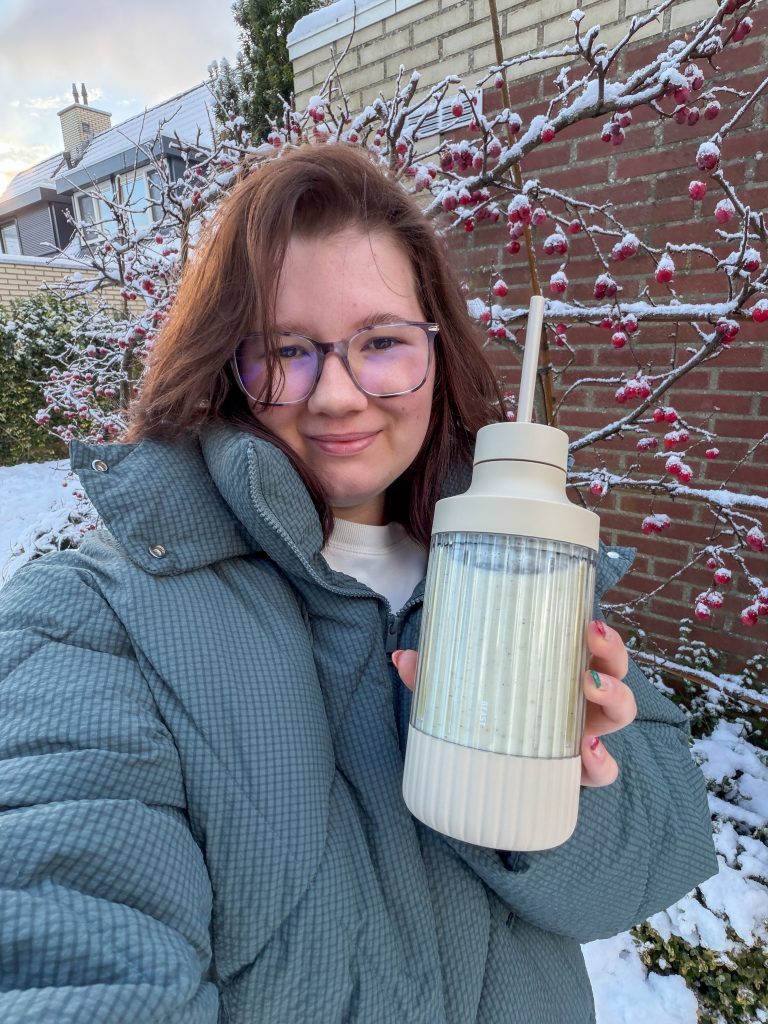Beaudine staat in de besneeuwde tuin en houdt een blenderfles vast tijdens een winterse ochtend.