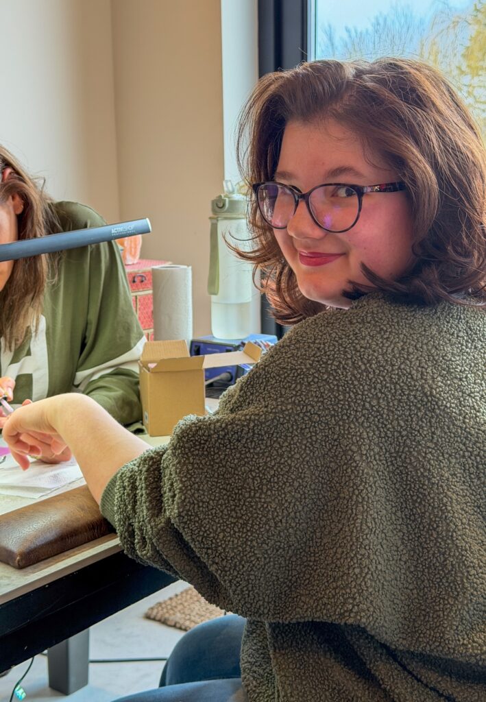 Beaudine zit bij de nagelstyliste en kijkt glimlachend in de camera