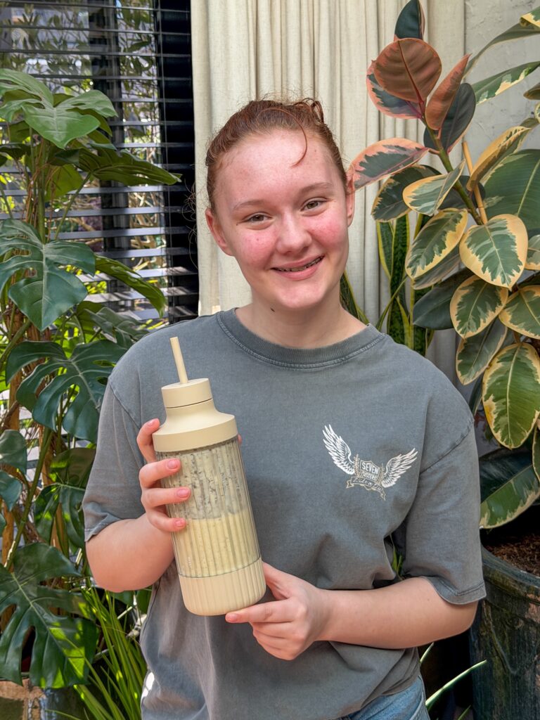 Amaranthe staat tussen de kamerplanten en laat een beige drinkbeker zien.