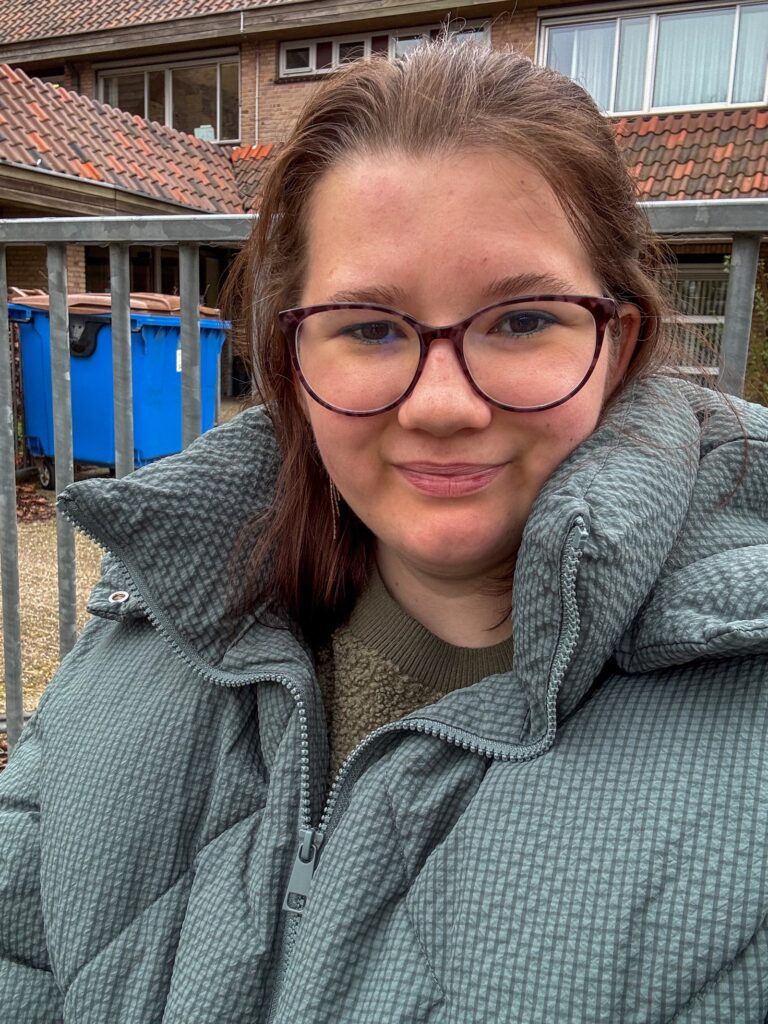 Beaudine in een groene jas buiten bij de kinderopvang, met gebouwen en blauwe containers op de achtergrond