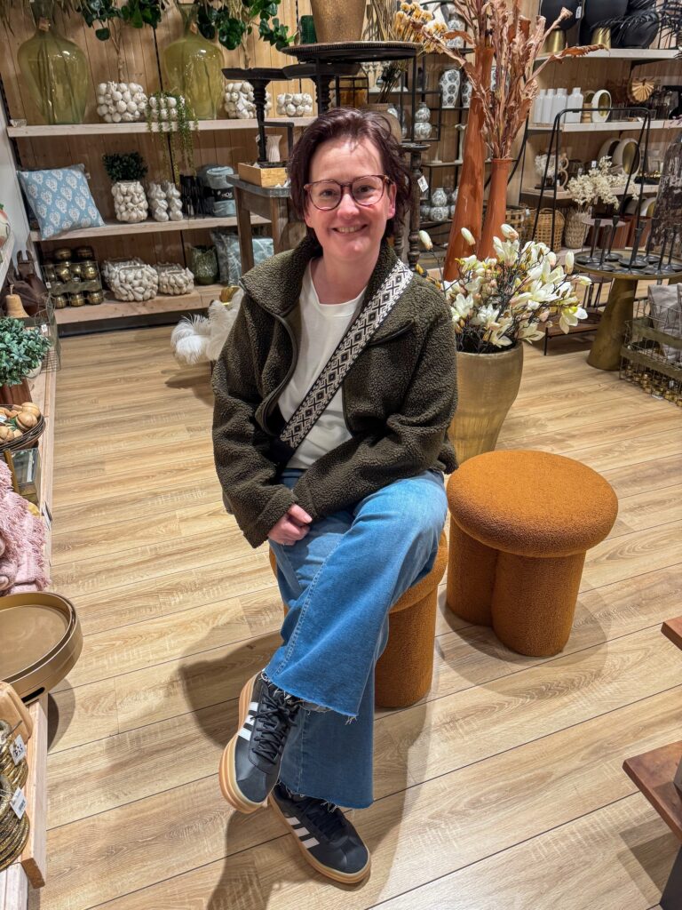 Marie Louise poseert ontspannen op een designkruk in een interieurwinkel met warme kleuren