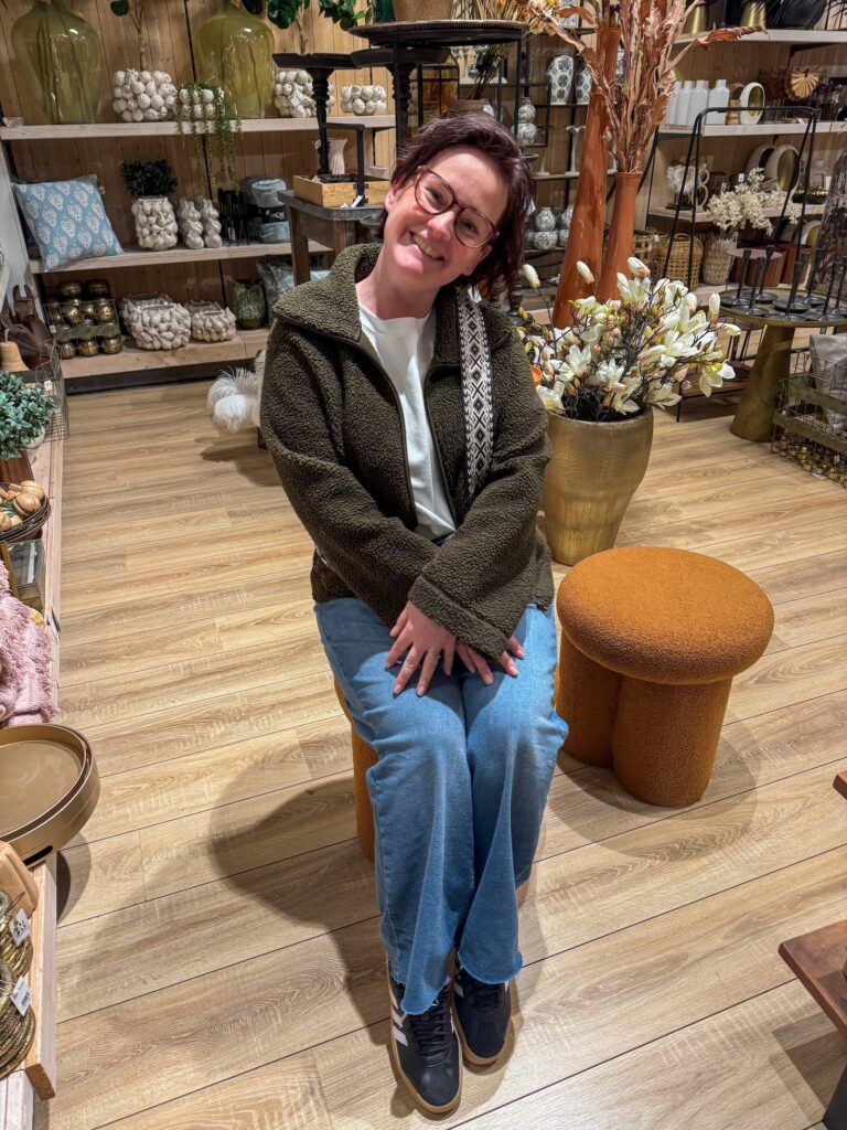 Marie Louise zit lachend op een ronde kruk in een sfeervolle woonwinkel vol vazen en decoratie