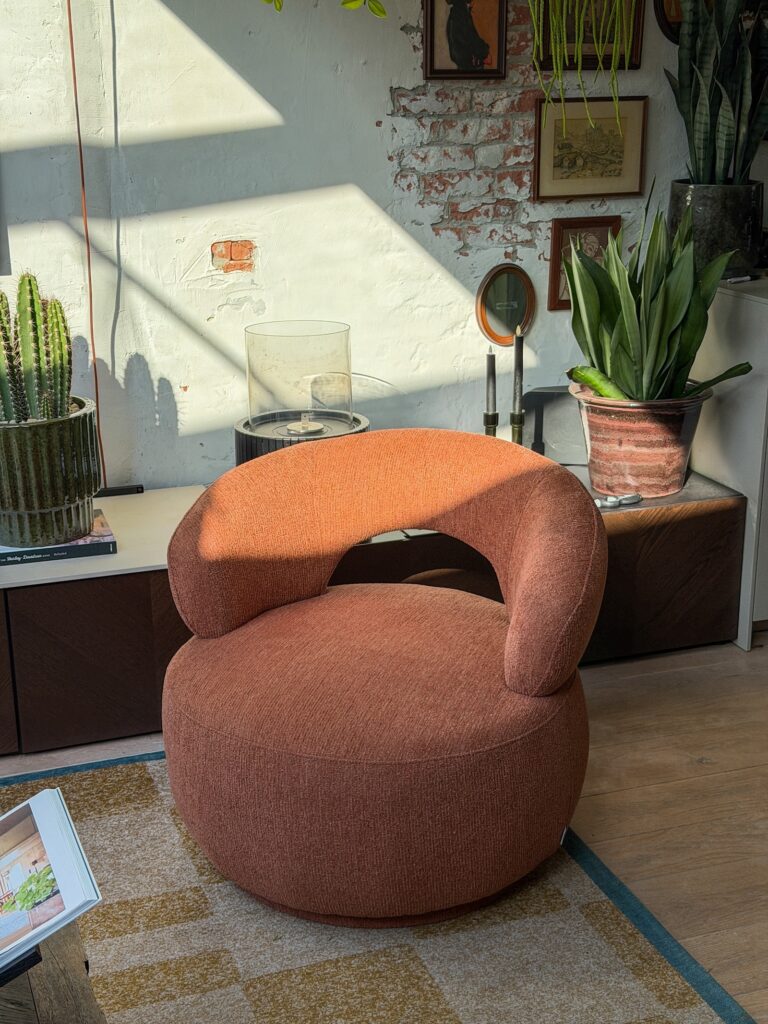 Oranje ronde fauteuil in onze woonkamer met planten, bakstenen muur en warm zonlicht.