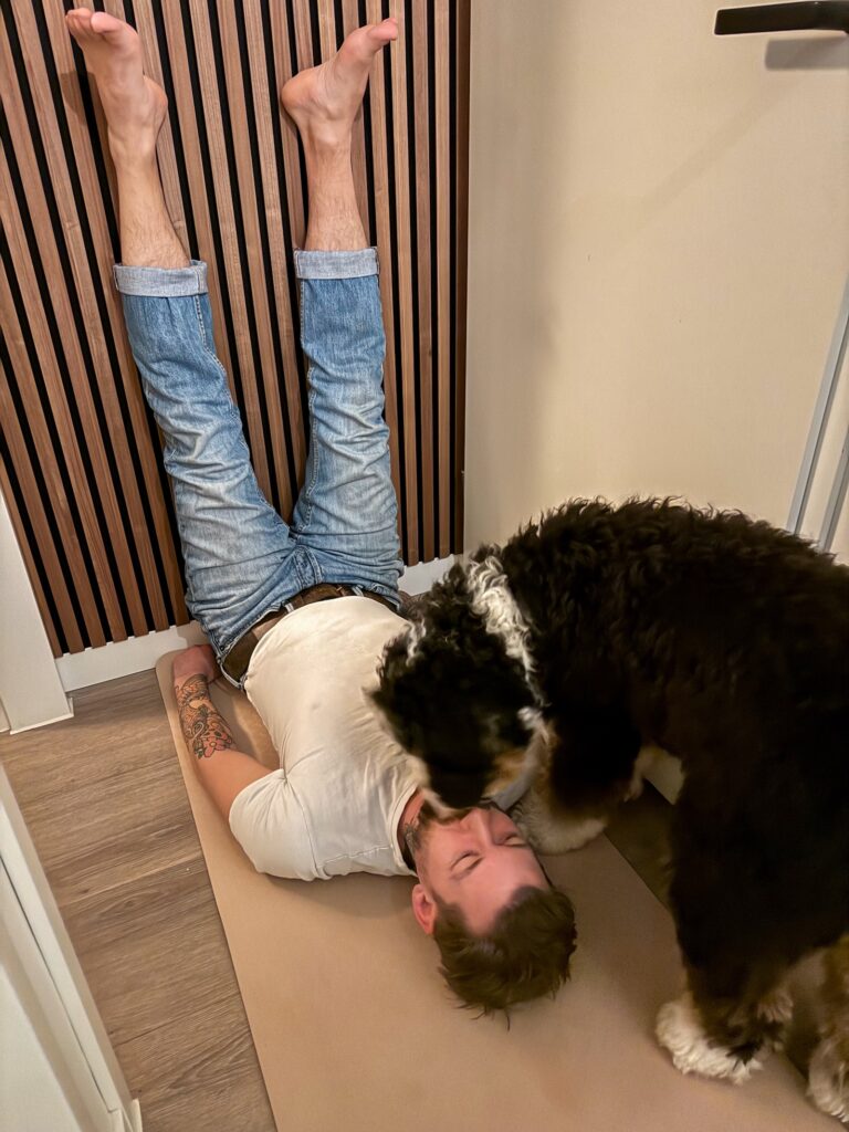 Jan willem ligt op yogamat met benen omhoog tegen de muur terwijl een Bernerdoodle nieuwsgierig over hem heen buigt