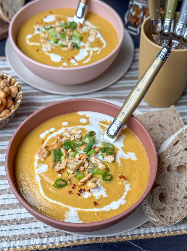 Kom met romige bloemkoolsoep met zoete aardappel en gele curry, gegarneerd met fijngesneden bosui en grof gehakte cashewnoten