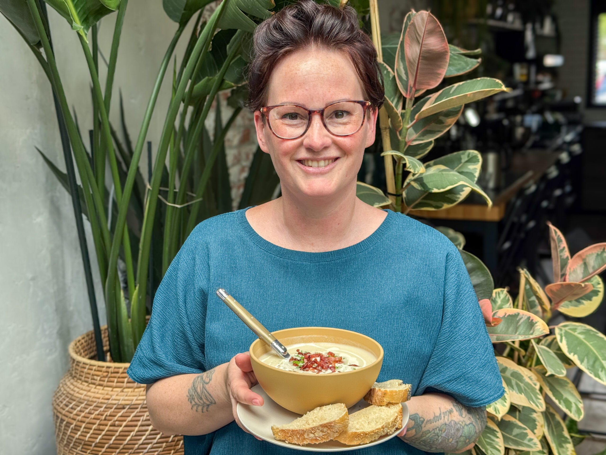 Marie Louise serveert romige bloemkoolsoep in gele kom met spekjes en brood, groene planten op achtergrond